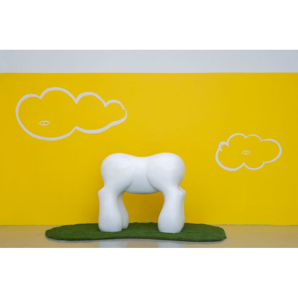 Installation photo showing a white, multi-legged sculpture positioned on green turf against a bright yellow wall. (Alt-text generated by AI)