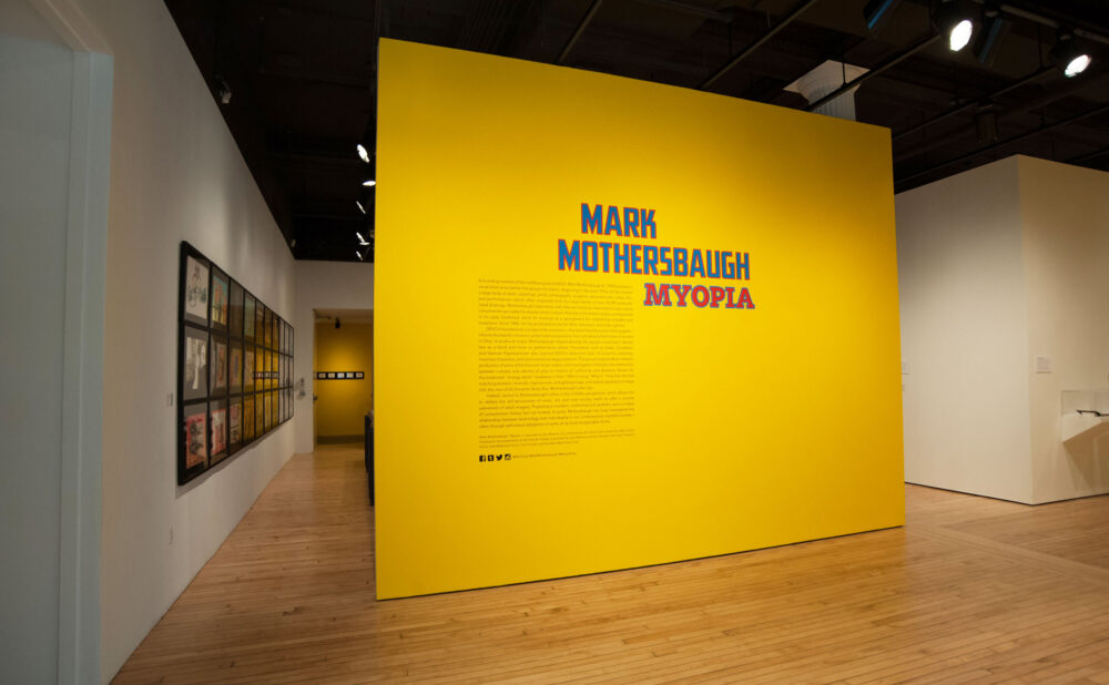 Perspective view of an exhibition entryway featuring a bold yellow accent wall printed with blue and red title lettering. (Alt-text generated by AI)