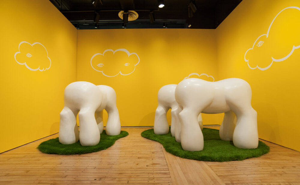 Bright yellow gallery alcove featuring two white, multi-legged animalistic sculptures standing on artificial grass patches. (Alt-text generated by AI)