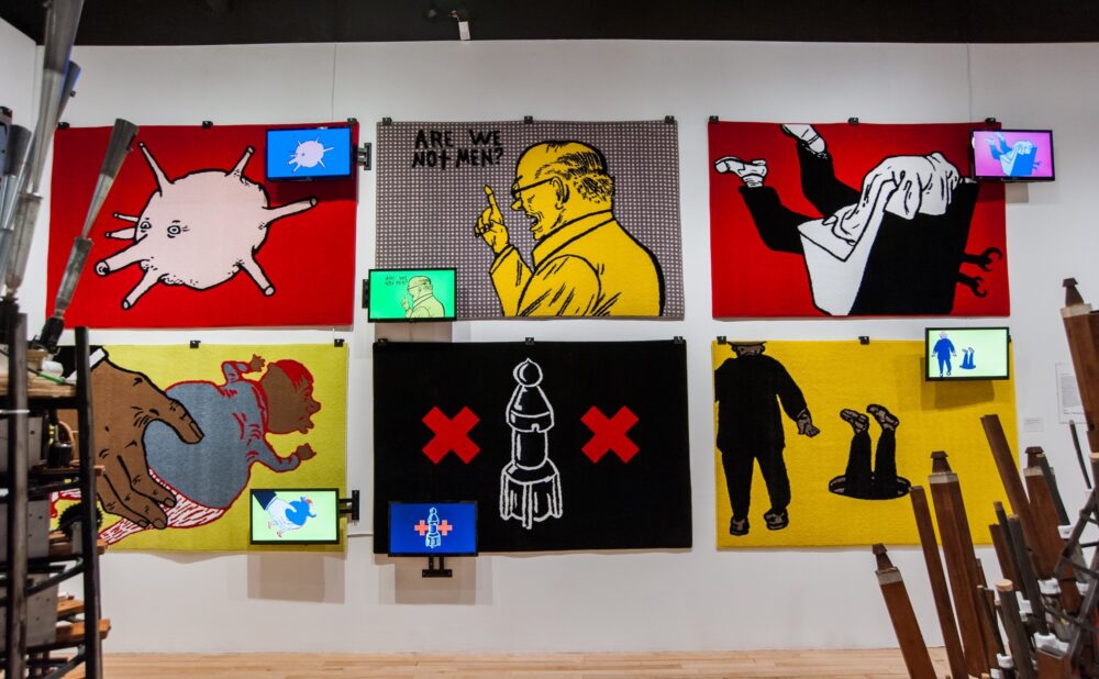 Exhibition wall featuring a dense grid layout of colorful graphic prints, video screens, and illustrations. (Alt-text generated by AI)