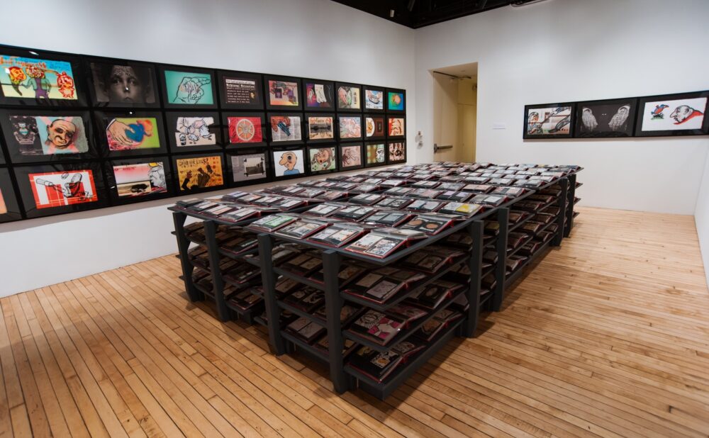 Open gallery view with a long, black, multirow display table positioned in front of framed artwork rows on the back wall. (Alt-text generated by AI)