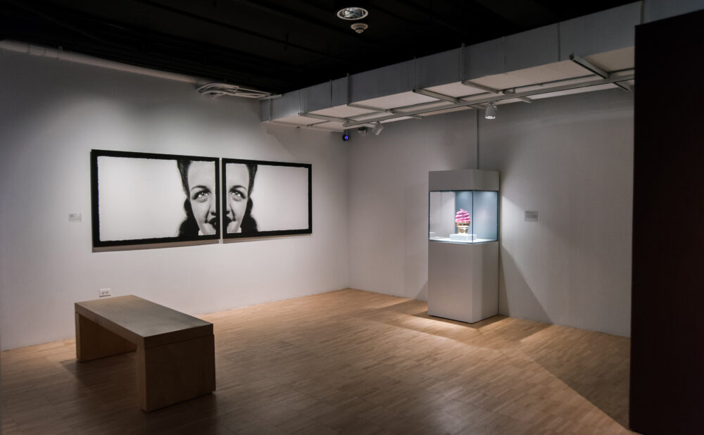 Dim gallery space with spotlight on a central tall display case, framed works mounted along the side walls, and a wooden bench. (Alt-text generated by AI)