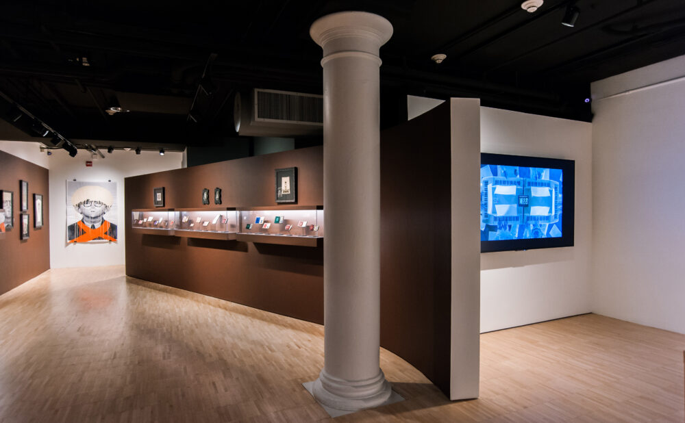 Wide view of the exhibition hall showing a central structural column, illuminated display cases on a brown wall, and a glowing video display. (Alt-text generated by AI)