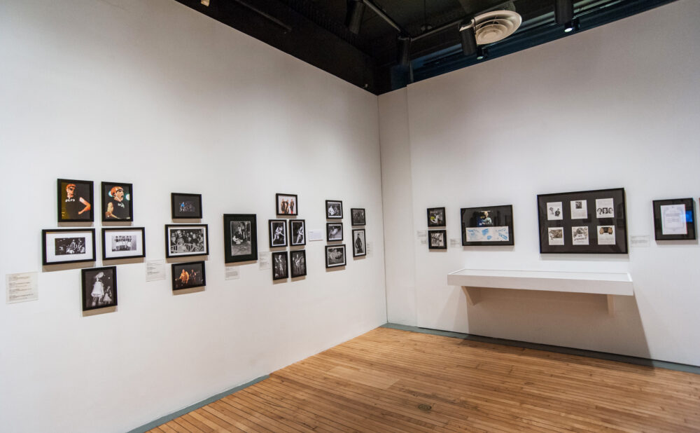 Gallery corner displaying rows of small framed artworks arranged horizontally on white walls above hardwood floors. (Alt-text generated by AI)