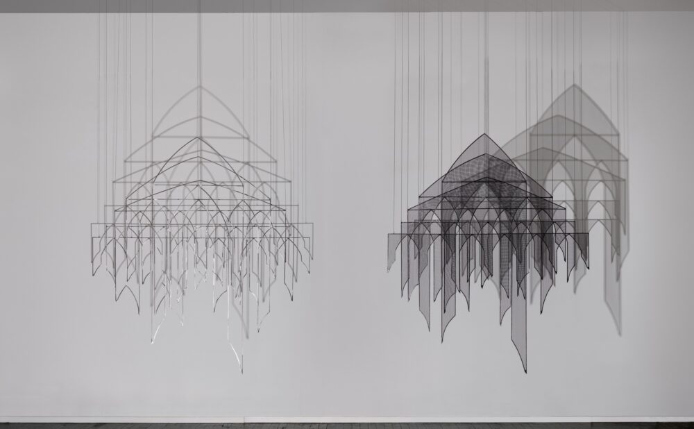 Two suspended geometric canopy sculptures with repeated arches, pointed hanging panels, vertical cords, and layered shadows on a gallery wall. Alt text generated by AI.