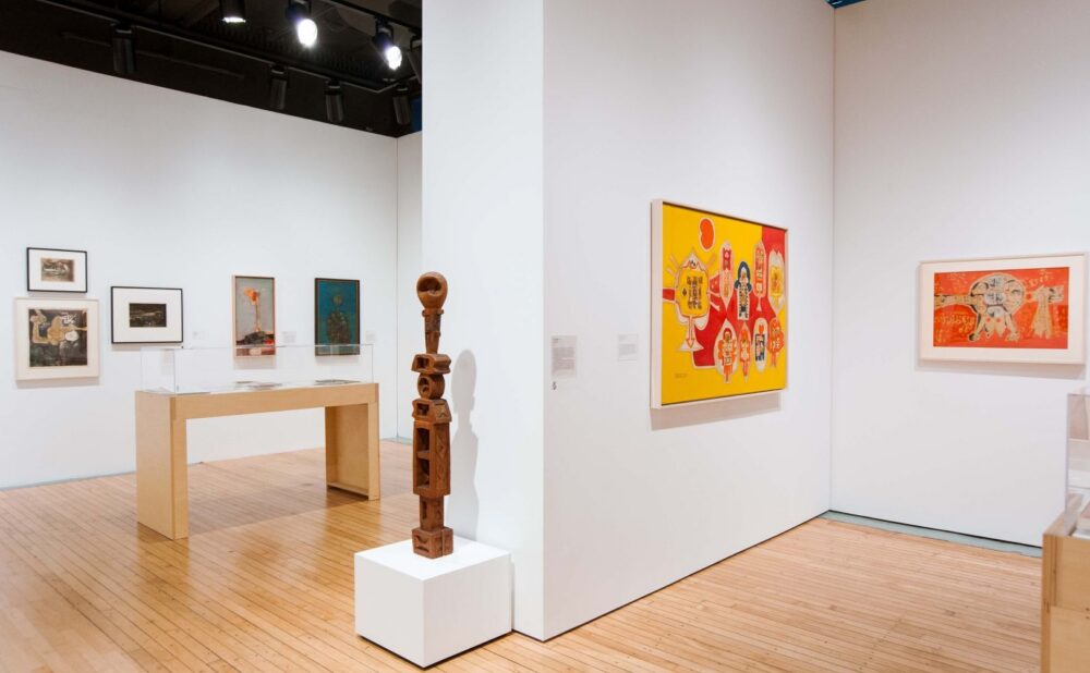 An art gallery with various artworks on white walls, including a large colorful painting, a smaller framed piece, and a wooden sculpture on a pedestal. Alt text generated by AI.