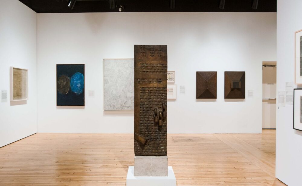 An art gallery featuring a central metal sculpture and various framed artworks on the walls, including abstract shapes and textured pieces, all set against wooden flooring. Alt text generated by AI.