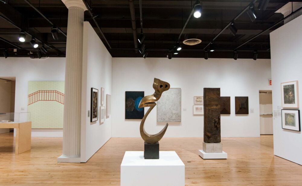 An art gallery displays a bronze abstract sculpture on a pedestal in the foreground, surrounded by various framed artworks on white walls, with a light wooden floor and a column in the background. Alt text generated by AI.