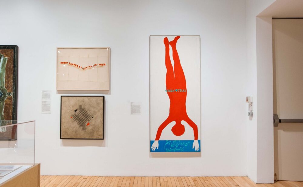 Gallery wall featuring three artworks: an abstract piece in earthy tones, a light artwork with orange lines, and a large red diving figure against a white background. Alt text generated by AI.