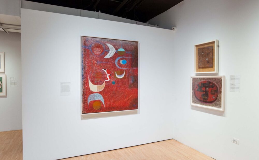 Interior of an art gallery with a large red abstract painting and two smaller framed artworks on a light-colored wall.