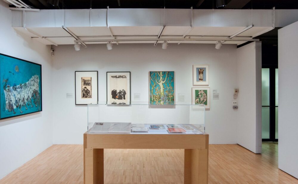 Interior of an art gallery featuring a large blue abstract painting, several framed artworks, and a glass display case with art publications. Alt text generated by AI.