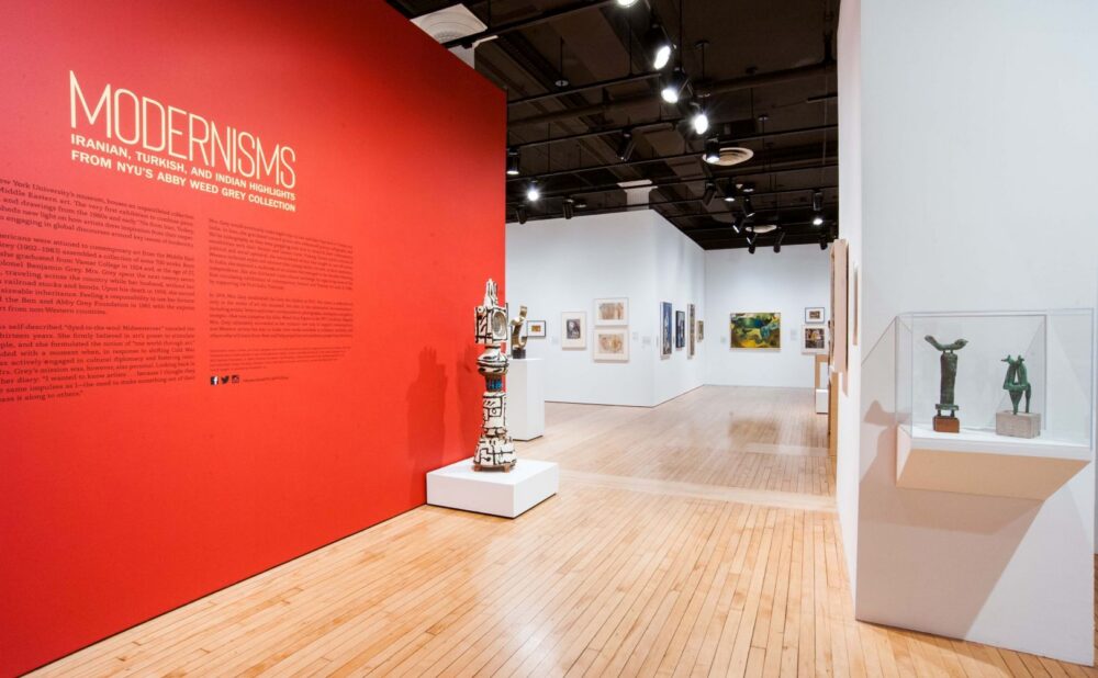 Interior of an art gallery featuring a red wall with exhibition text, a sculpture in the foreground, framed artworks on the walls, and a glass display case with sculptures. Alt text generated by AI.