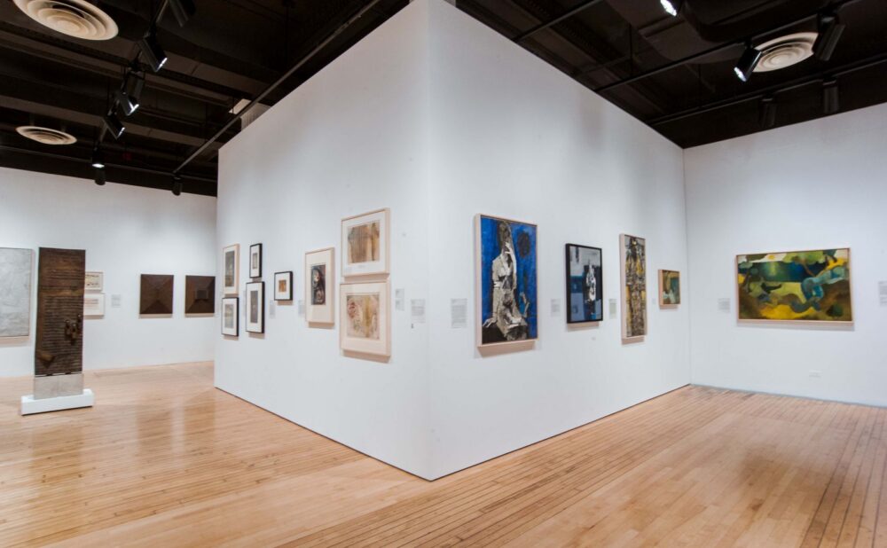 An art gallery with two white walls featuring a variety of framed artworks, a sculpture on a pedestal, and light wooden flooring. Alt text generated by AI.