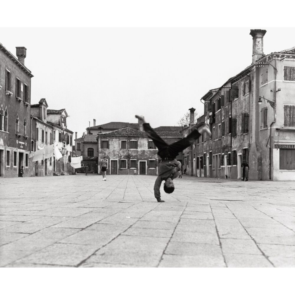 Black-and-white photograph of a person performing a handstand in an open town square lined with historic buildings. (Alt-text generated by AI)