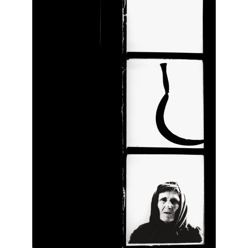 Vertical high-contrast photograph featuring an sickle shape above and an elderly figure below. (Alt-text generated by AI)
