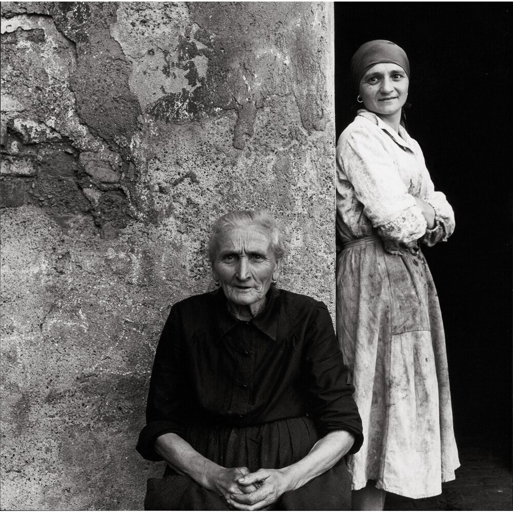Black-and-white portrait showing an elderly woman seated beside a standing younger woman outdoors against a stone wall. (Alt-text generated by AI)