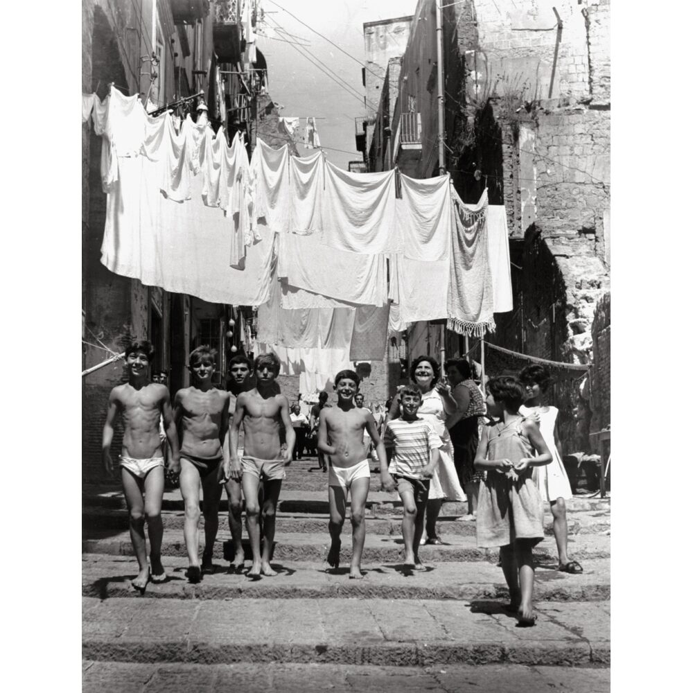 Black-and-white street scene showing children playing in a narrow alleyway underneath hanging laundry lines. (Alt-text generated by AI)