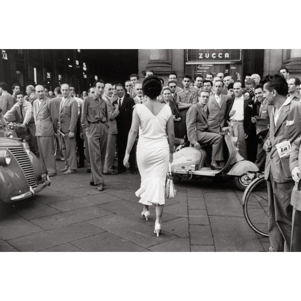 Black-and-white photograph capturing a woman walking down a crowded street while surrounding onlookers turn to watch her pass. (Alt-text generated by AI)
