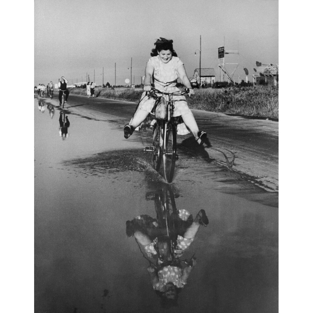 Black-and-white photo of a person riding a bicycle along a wet road with reflections visible on the ground. (Alt-text generated by AI)