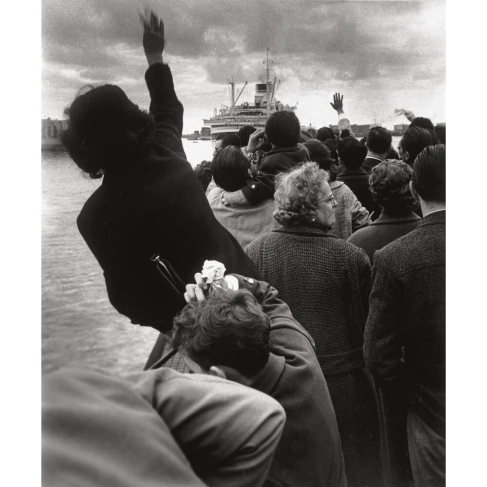 Black-and-white photograph depicting a crowded group of people outdoors waving towards the distance. (Alt-text generated by AI)