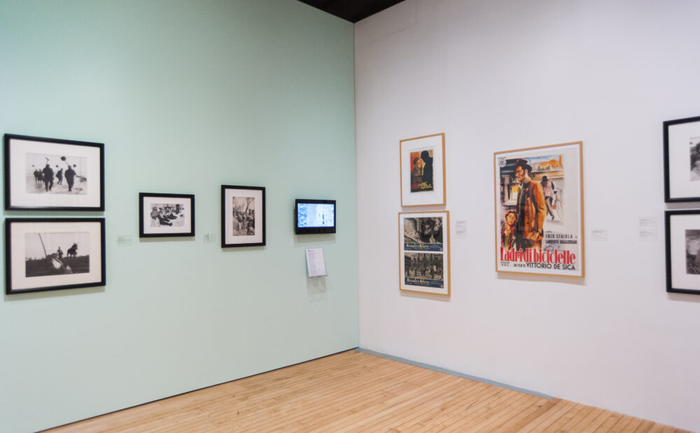 Exhibition space showing a mint-green wall on the left meet a white wall on the right mounted with framed pictures and posters. (Alt-text generated by AI)