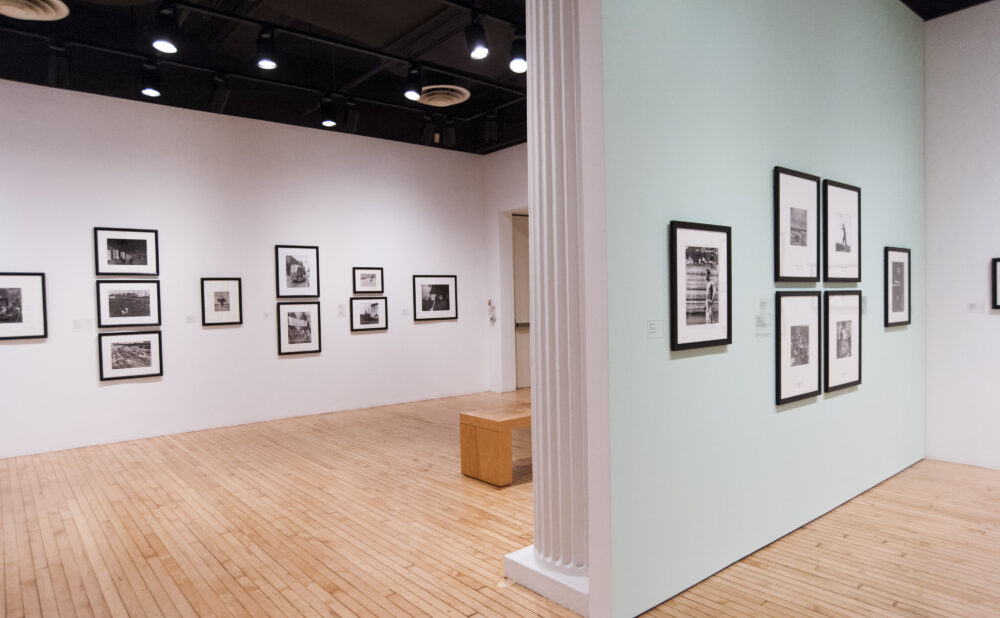 View of a gallery corridor showing black-and-white framed photographs mounted along pale green and white walls. (Alt-text generated by AI)