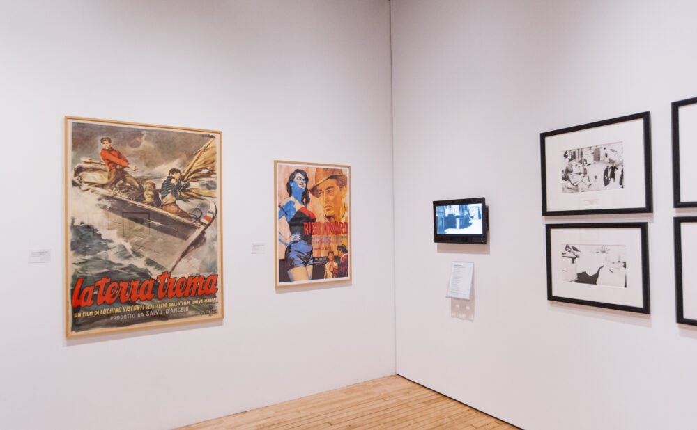 Corner of a gallery room displaying vintage Italian film posters on the left and framed black-and-white prints on the right. (Alt-text generated by AI)