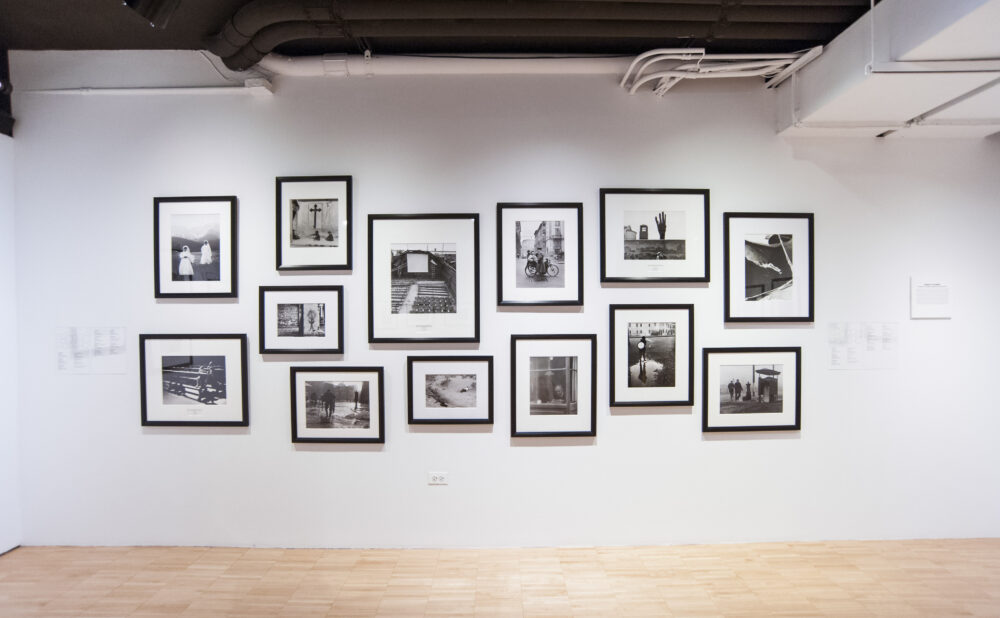 White gallery wall featuring a grid layout of small black-and-white framed photographs. (Alt-text generated by AI)