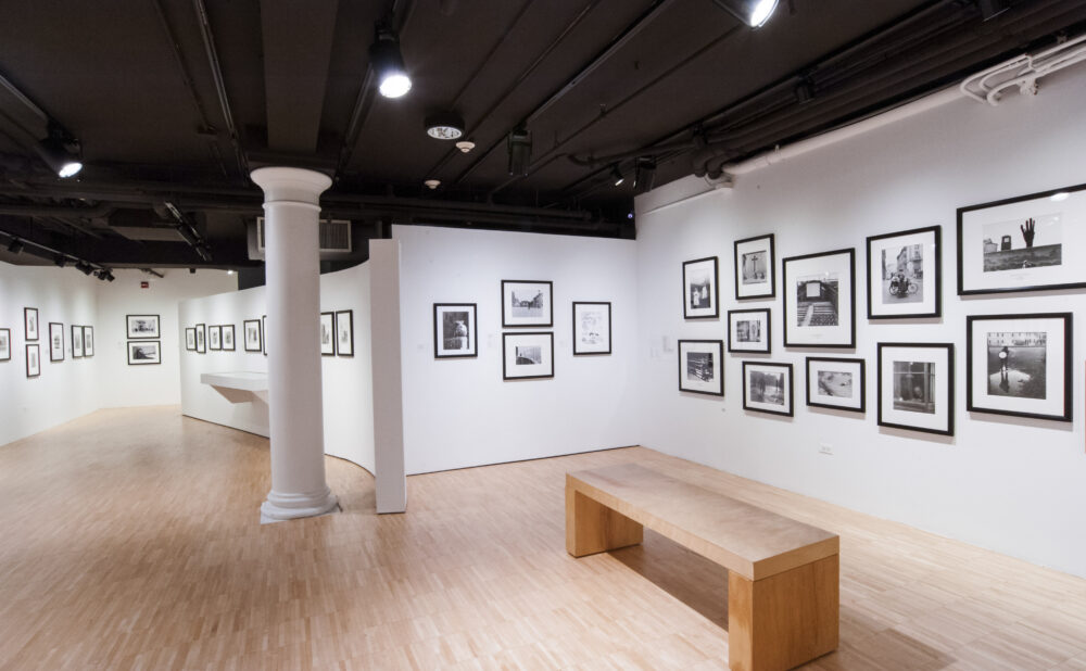 Wide gallery view featuring rows of framed black-and-white photographs, a structural pillar, and a wooden bench. (Alt-text generated by AI)