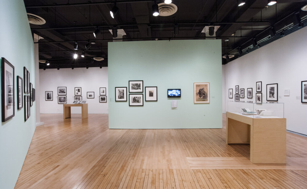 Open exhibition hall showing mint-green partition walls, framed photographs, and a wooden display case. (Alt-text generated by AI)