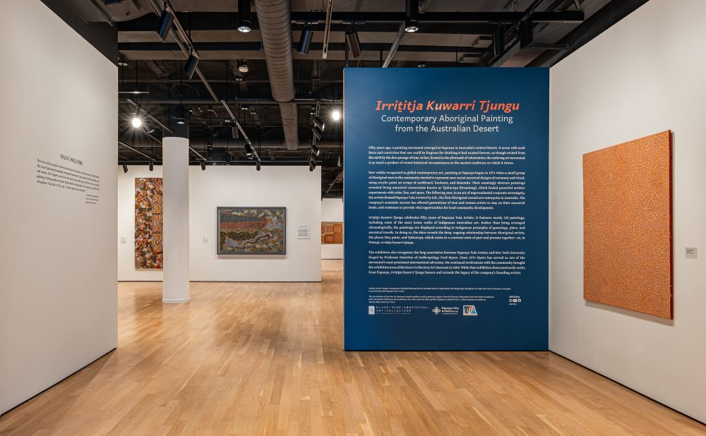 Gallery featuring contemporary Aboriginal paintings with an informational blue panel on the exhibition.