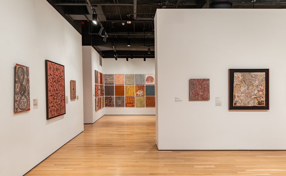 Art gallery with wooden floors and white walls displaying several Aboriginal paintings with intricate patterns and warm colors.