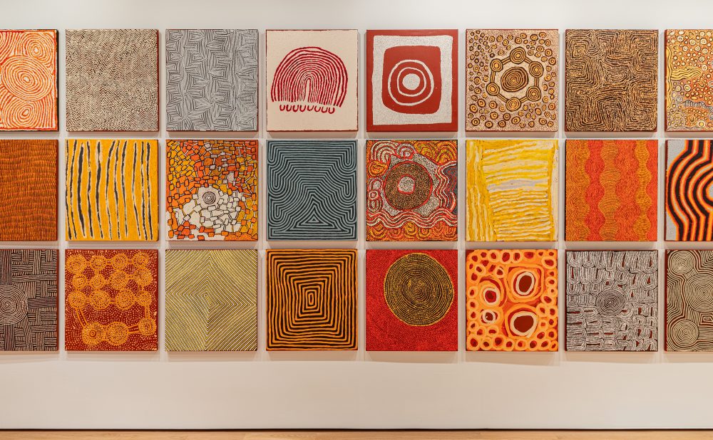 Art gallery with wooden floors and white walls displaying a grid of square Aboriginal paintings with intricate patterns and warm colors.