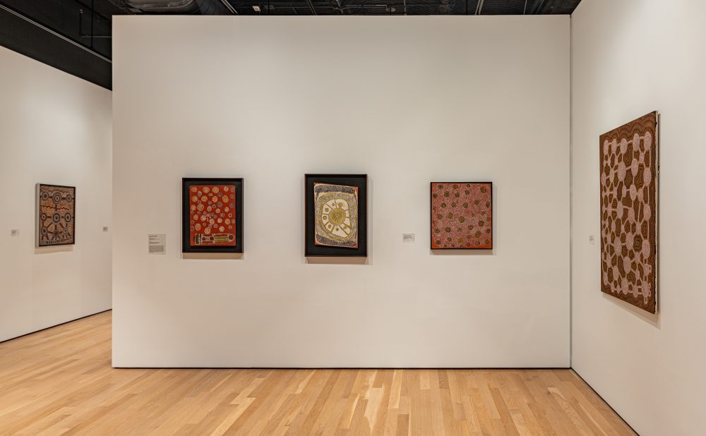 Art gallery with wooden floors and white walls displaying several Aboriginal paintings with intricate patterns and warm colors.