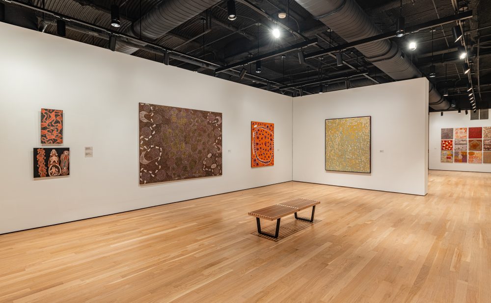 Art gallery with wooden floors and white walls displaying several Aboriginal paintings with intricate patterns and warm colors.