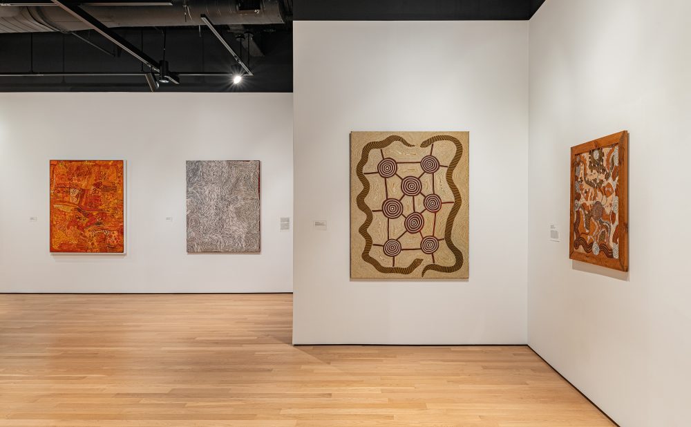 Art gallery with wooden floors and white walls displaying four Aboriginal paintings with intricate patterns and warm colors.