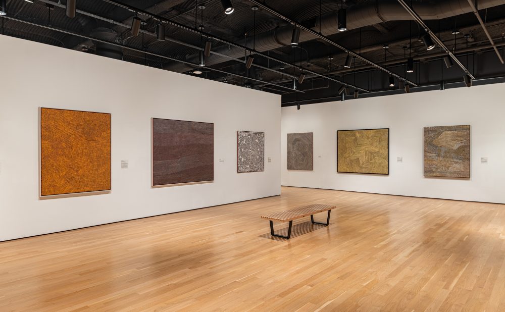 Art gallery with wooden floors and white walls displaying several Aboriginal paintings with intricate patterns and warm colors.