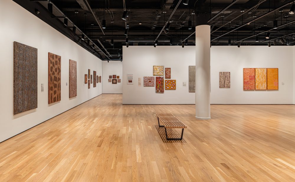 Art gallery with wooden floors and white walls displaying several Aboriginal paintings with intricate patterns and warm colors.