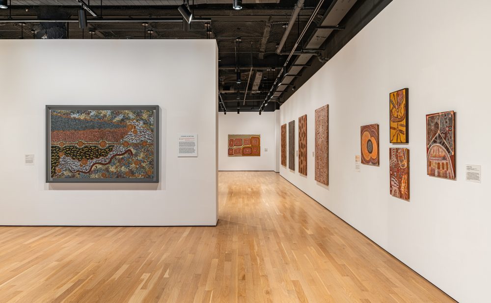 Art gallery with wooden floors and white walls displaying several Aboriginal paintings with intricate patterns and warm colors.