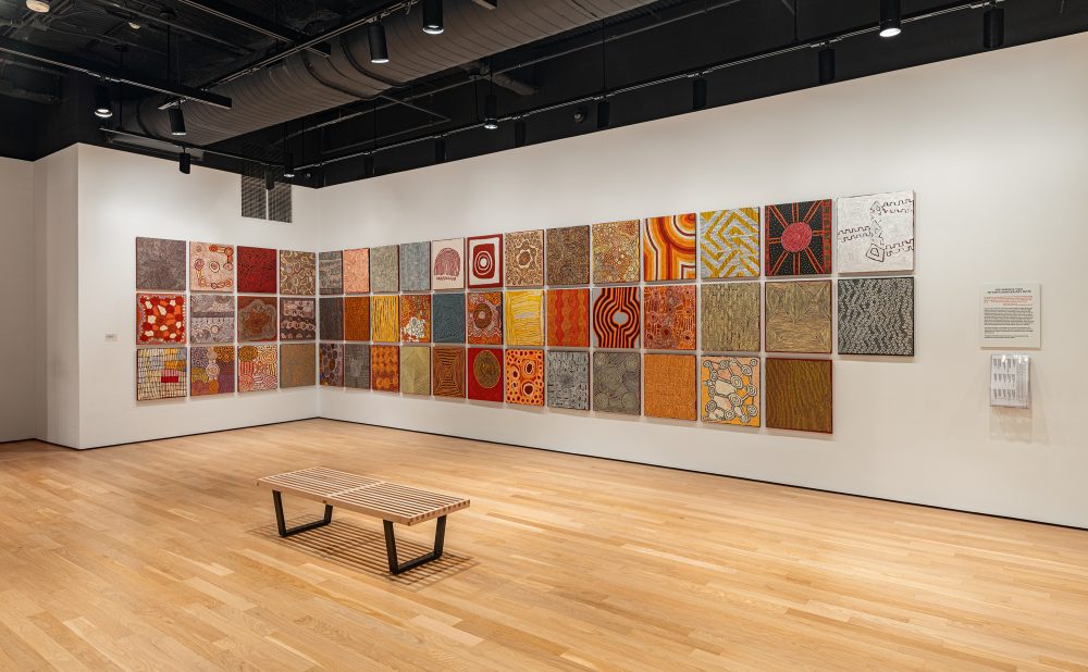 Art gallery with wooden floors and white walls displaying a grid of Aboriginal paintings with intricate patterns and warm colors.