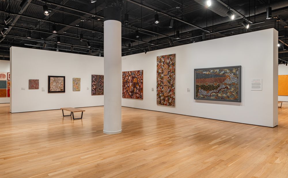 Art gallery with wooden floors and white walls displaying several Aboriginal paintings with intricate patterns and warm colors.