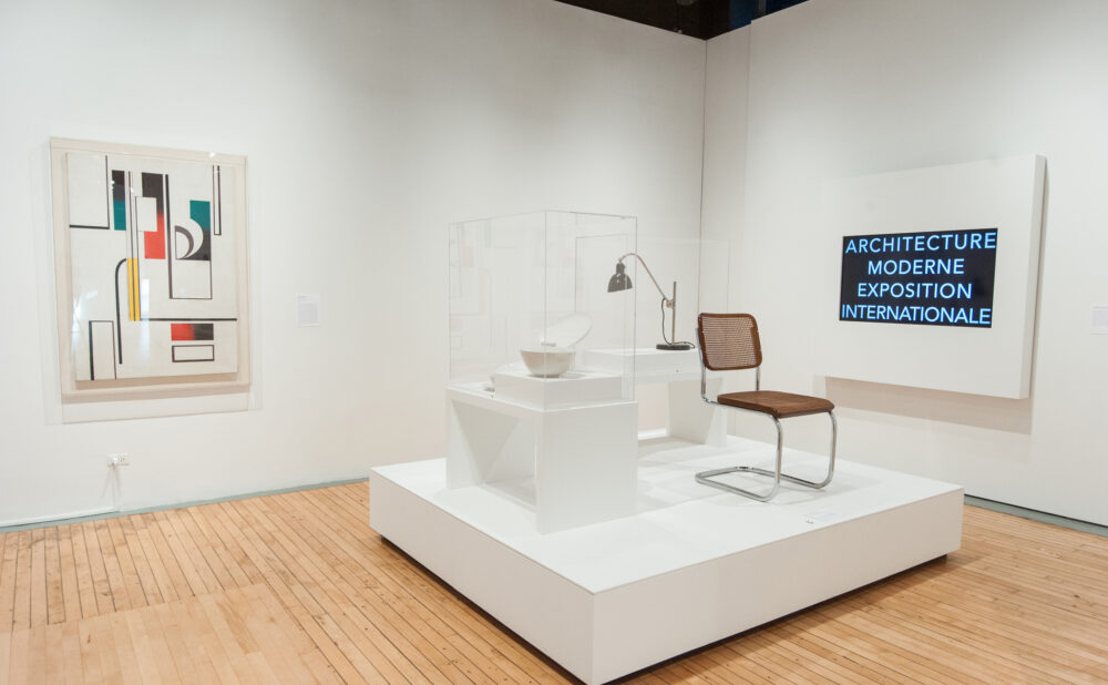 Corner exhibition view featuring modern chairs displayed on a low white platform alongside framed architectural prints on the walls. (Alt-text generated by AI)
