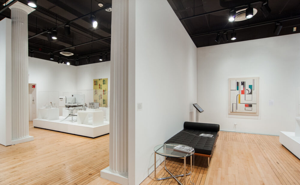 Wide gallery perspective showing polished wood floors, architectural columns, and black-and-silver lounge furniture in the foreground. (Alt-text generated by AI)