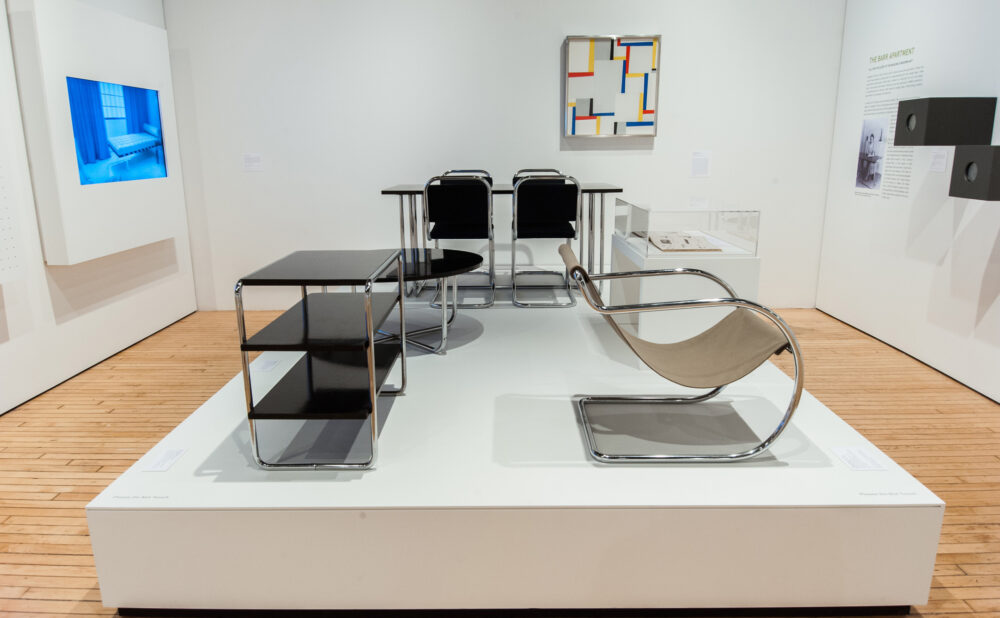 Exhibition platform displaying iconic tubular steel chairs and glass side tables against a white gallery wall. (Alt-text generated by AI)