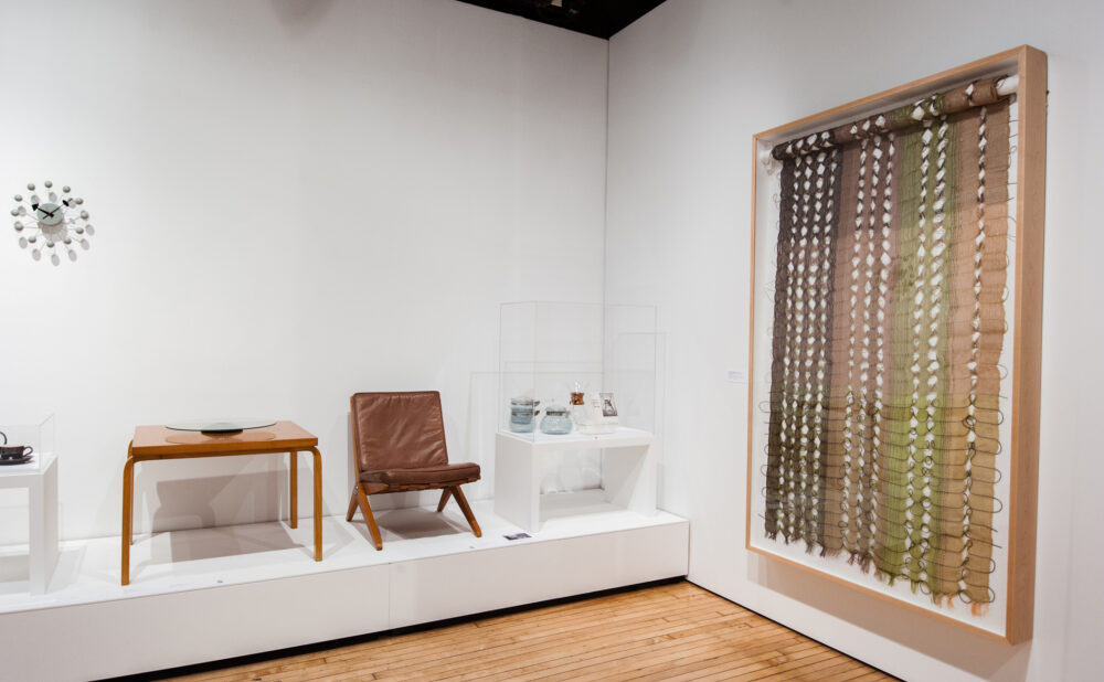 Gallery wall section featuring wooden modern chairs on a platform, a small table, and a large textured wall hanging. (Alt-text generated by AI)