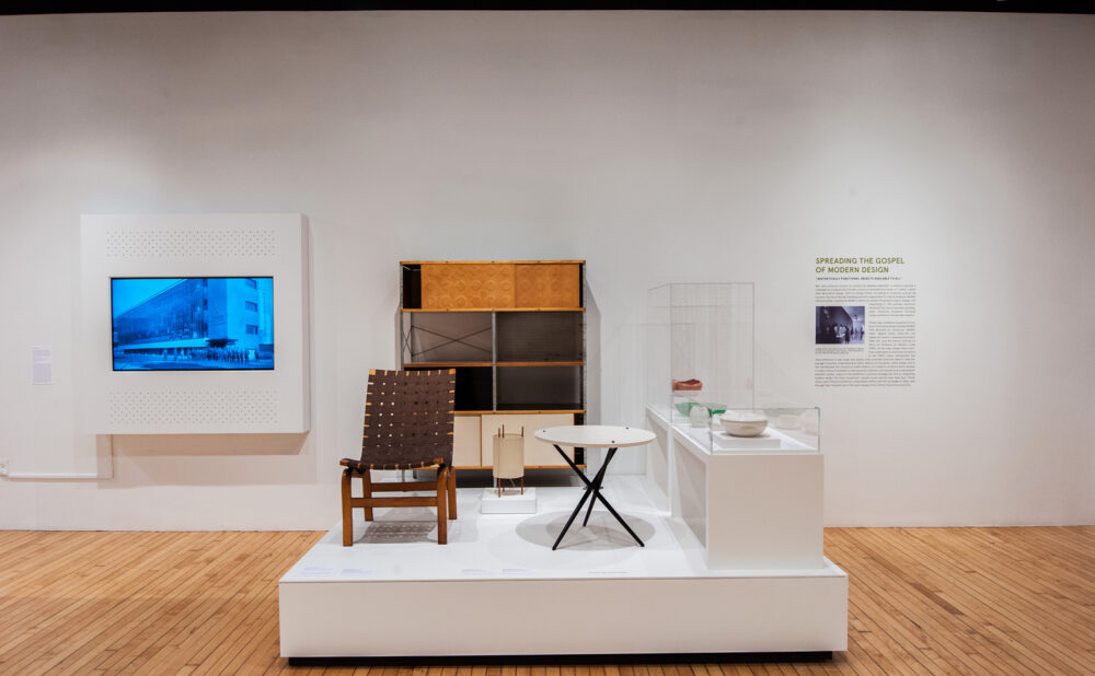 Display area showing mid-century chairs, small tables, and a cabinet set against white walls with mounted artwork. (Alt-text generated by AI)