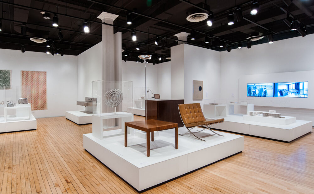Open gallery view with a central raised platform holding a mid-century wooden lounge chair and coffee table. (Alt-text generated by AI)