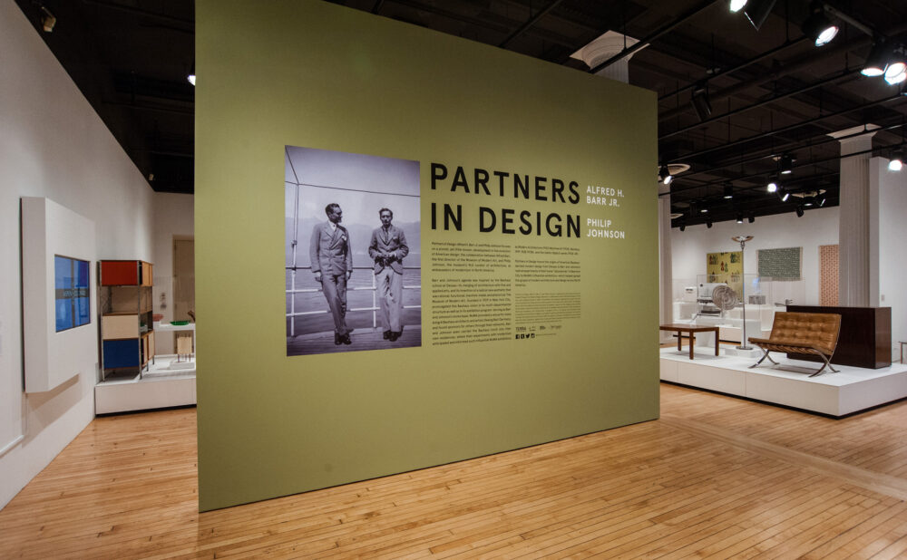 Gallery entrance area featuring an olive-green wall with the exhibition title "PARTNERS IN DESIGN" printed above a large archival photoreproduction. (Alt-text generated by AI)