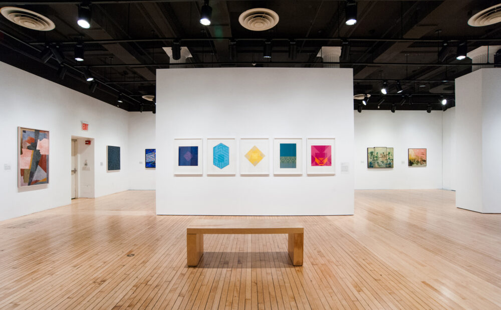 An art gallery with abstract geometric artworks on display and a wooden bench in the center.