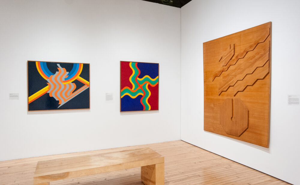 Gallery room with three abstract artworks, a wooden bench and wood flooring.